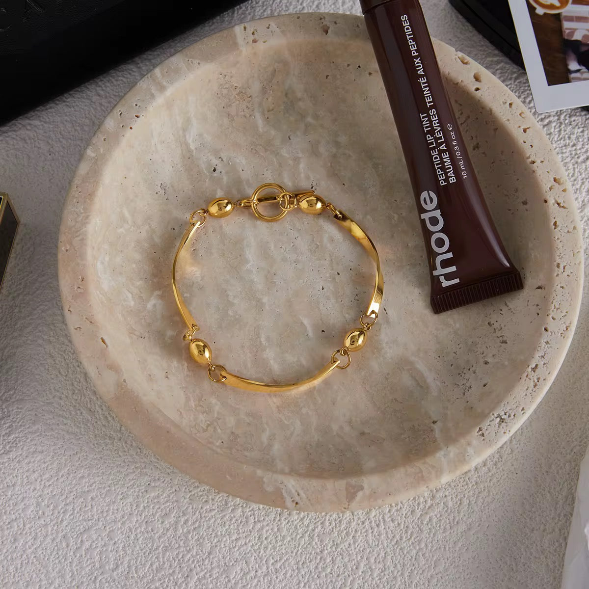 Gold bracelet on a stone surface with a tube of lip balm labeled rhode.