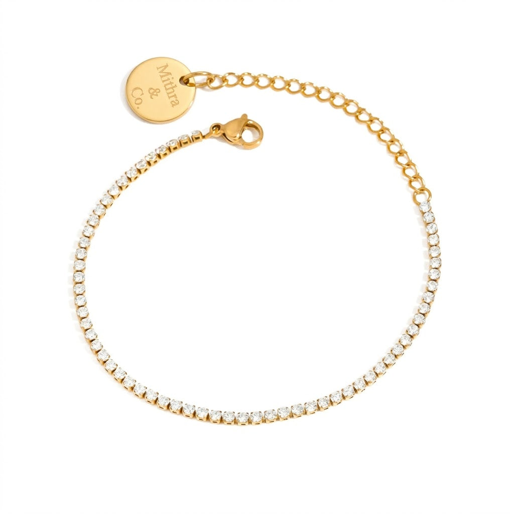 A delicate gold chain bracelet with a row of small clear stones, a lobster clasp, and an adjustable extender chain featuring a round engraved gold tag that reads 'Mila & Co'.