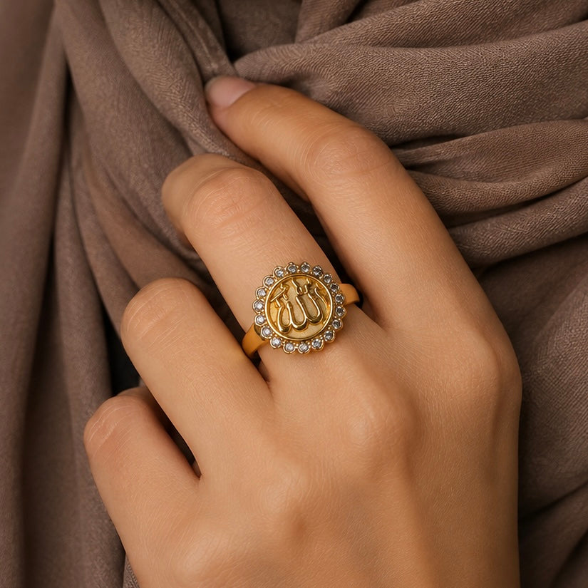Allah medallion ring. A gold arabic signet ring with a central Allah name written in arabic calligraphy and a halo of small clear stones, worn on a person's finger against draped brown fabric.
