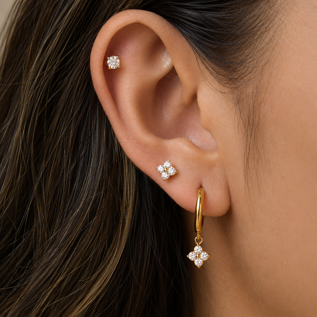 Close-up of a person's ear wearing three gold ear piercings: a small clvoer stud at the lobe, a small clover cluster stud above, and a gold hoop with a dangling clover-shaped cluster earring.