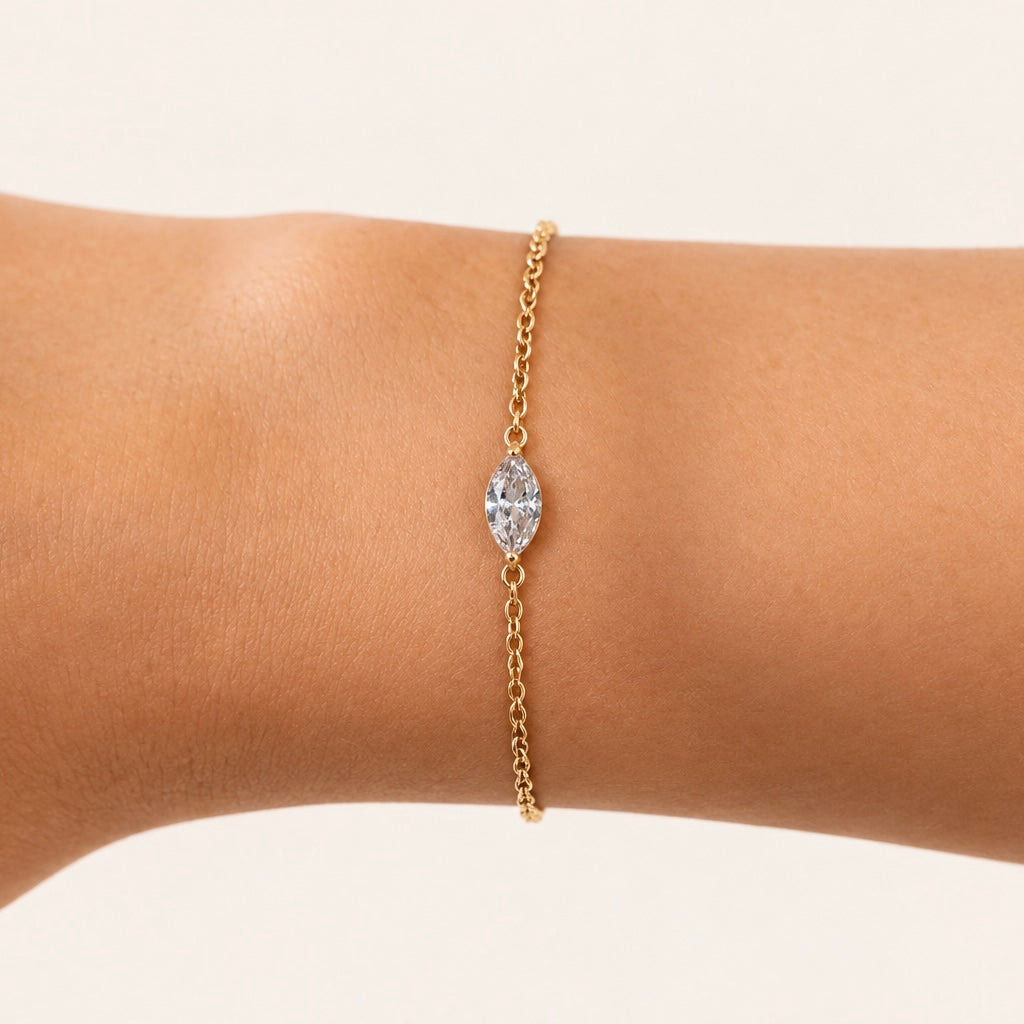 A delicate gold plated Leaf Charm chain bracelet with a marquise-cut clear gemstone centerpiece, shown worn on a person's wrist against a soft cream background.