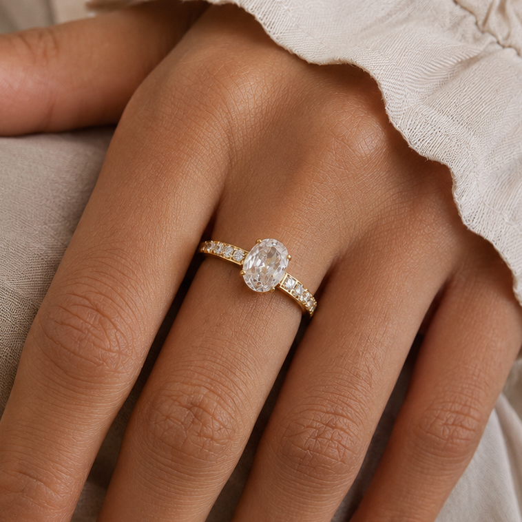 A close-up of a hand wearing a gold plated oval pave ring with a large oval clear center stone and small side stones, set against soft neutral fabric.