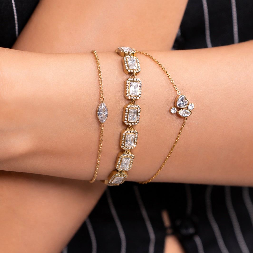 Three gold-plated bracelets with clear gemstones are shown worn on a person's wrist. The center bracelet features a row of rectangular clear stones, the left bracelet has a single marquise-shaped clear stone, and the right bracelet has a small cluster of clear stones with a heart-shaped accent.