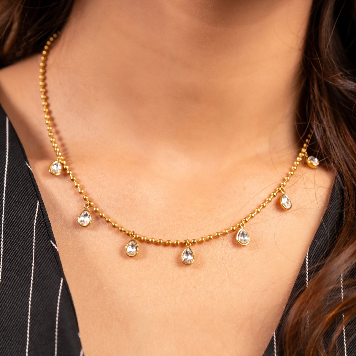 A close-up of a woman wearing a delicate gold necklace with five small teardrop-shaped clear stones hanging from a beaded gold chain.