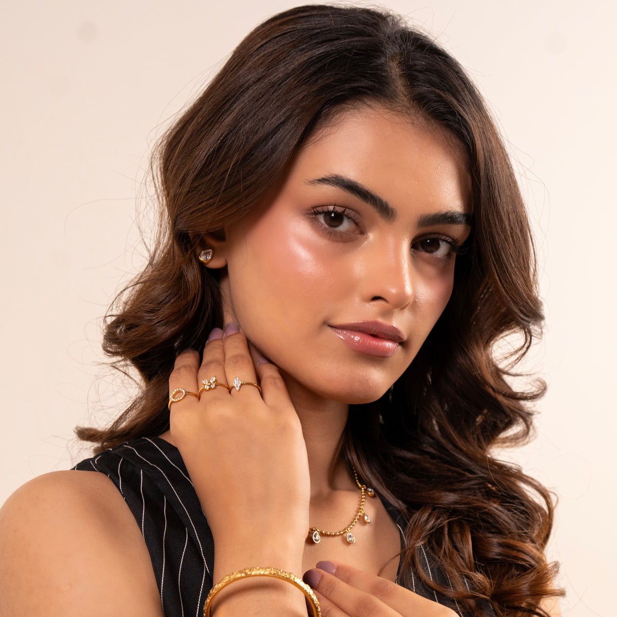Hana Soiree: A close-up portrait of a woman with long wavy brown hair, wearing gold jewelry including a delicate gold necklace, a gold ring, and a gold bracelet, set against a plain light beige background.
