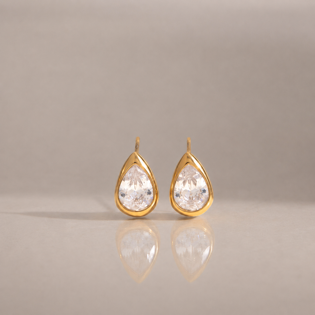 A pair of gold teardrop-shaped stud earrings with clear pear-cut gemstones, displayed on a neutral beige reflective surface.