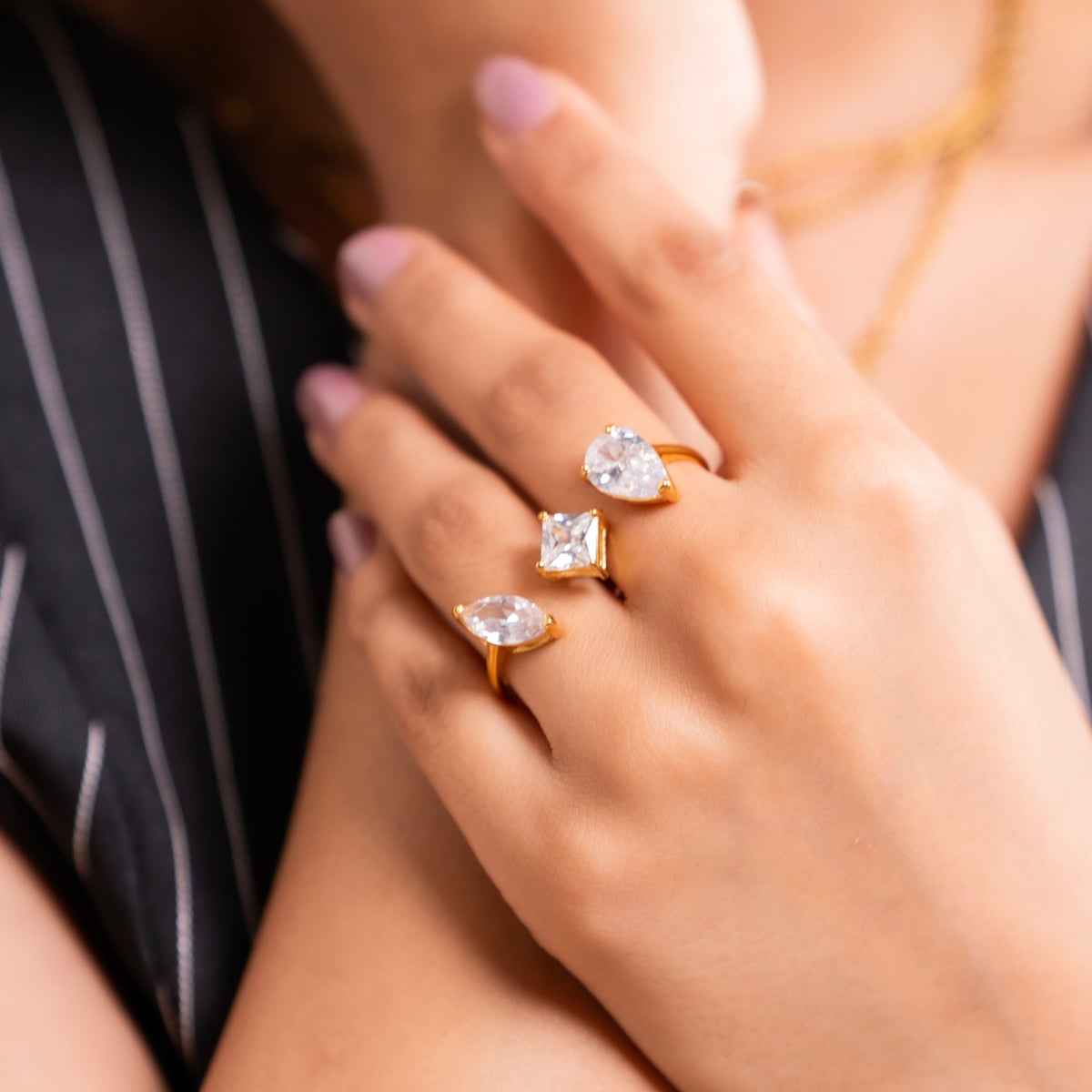 Three gold rings with clear gemstones in marquise, square, and pear shapes, worn on a person's fingers. The person is wearing a black and white pinstriped top.