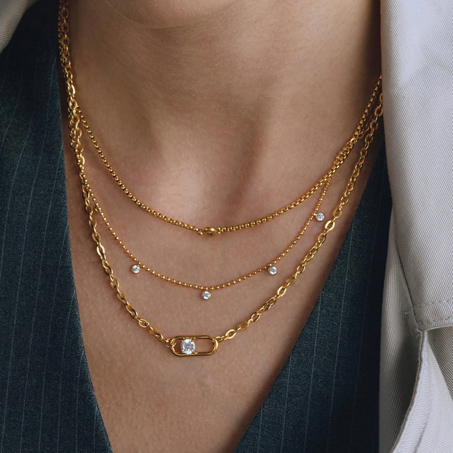 A close-up of a person's neck and upper chest wearing a 3 layered gold plated chain necklace set. The set includes a delicate chain, a twisted chain with small clear stones, and a safety-pin style chain with a clear central stone.