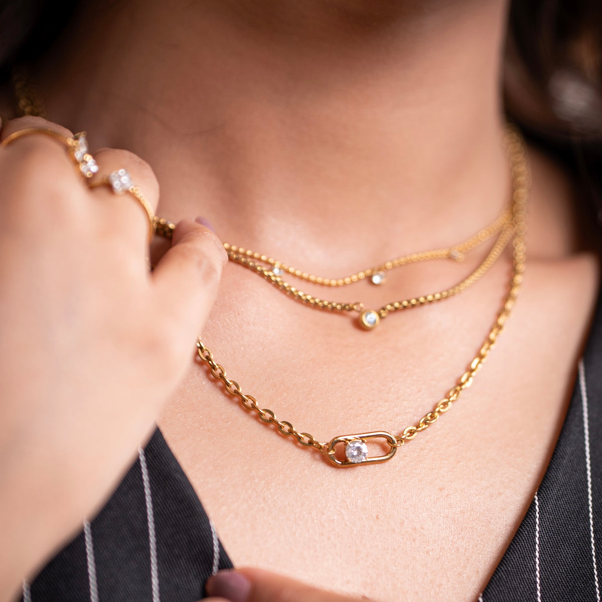 A close-up lifestyle shot of a model wearing a gold plated triple layered chain necklace set, featuring a delicate chain with a small gold bead and a safety-pin style chain with a clear rhinestone clasp, along with a matching gold ring on her finger.