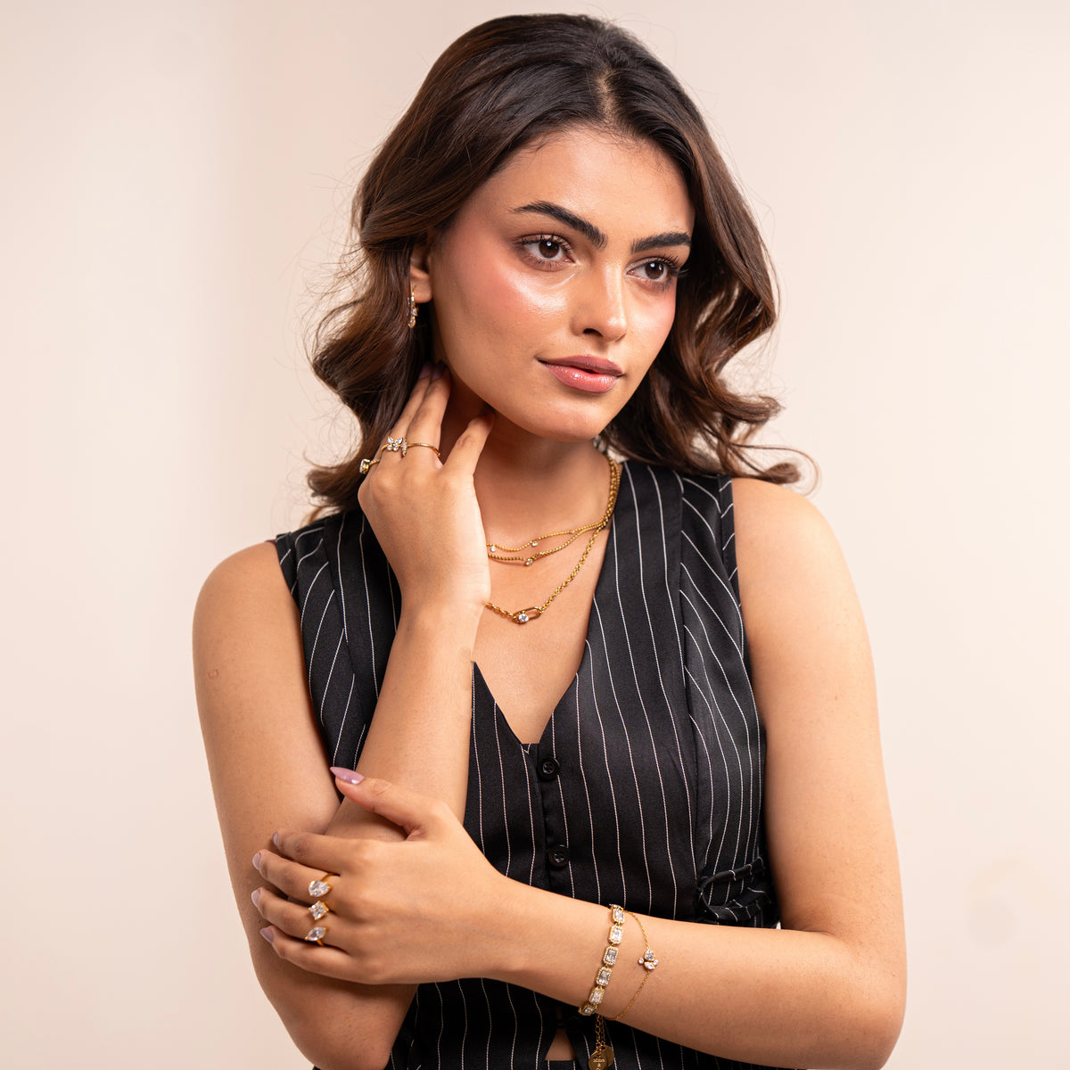 A woman with wavy brown hair models gold jewelry, including triple layer chain necklace, rings, and a bracelet, while wearing a black and white pinstripe sleeveless top against a plain beige background.