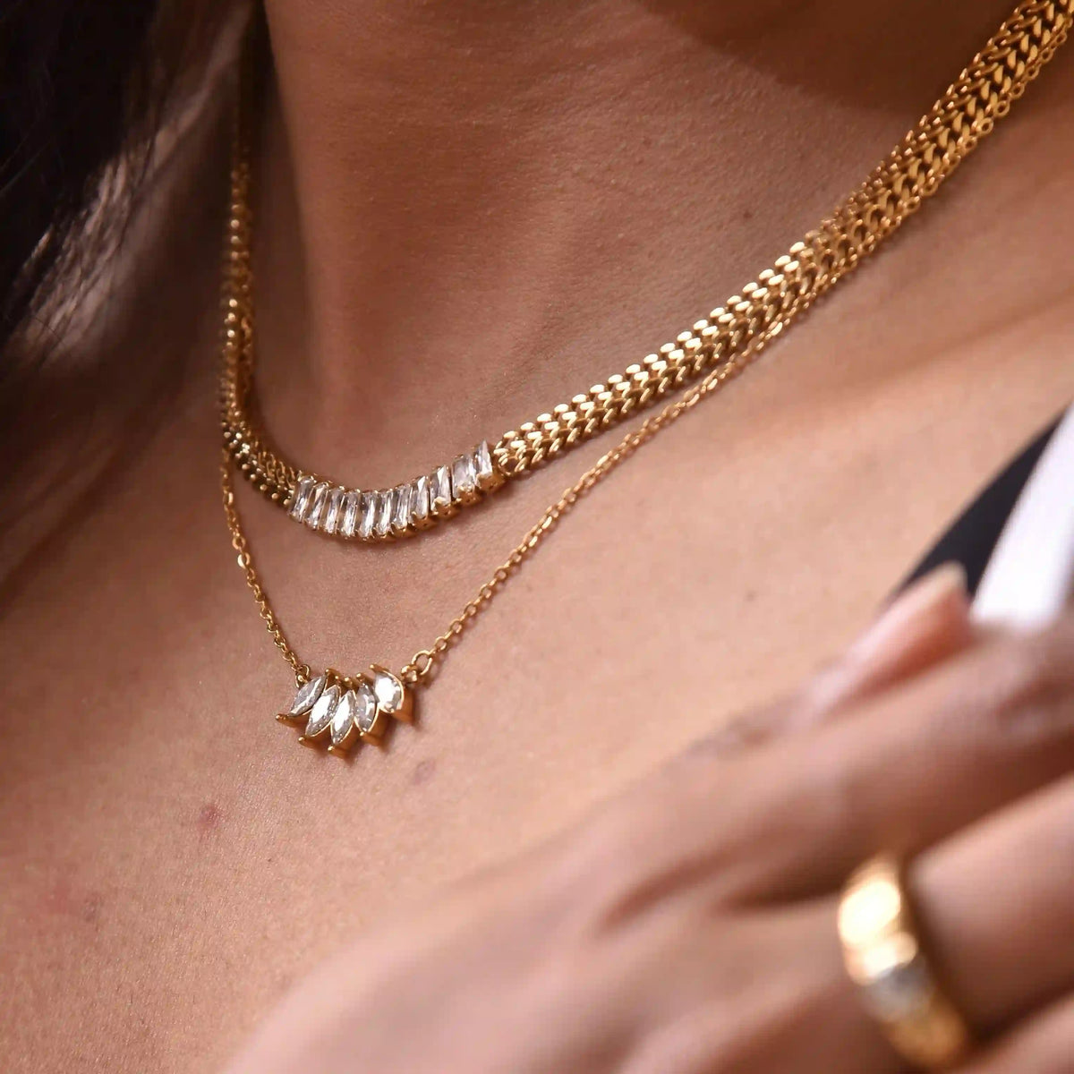 Close-up of a person wearing two gold plated necklaces, lilly pendant, and cleopatra neckchain with a ring on a neutral background