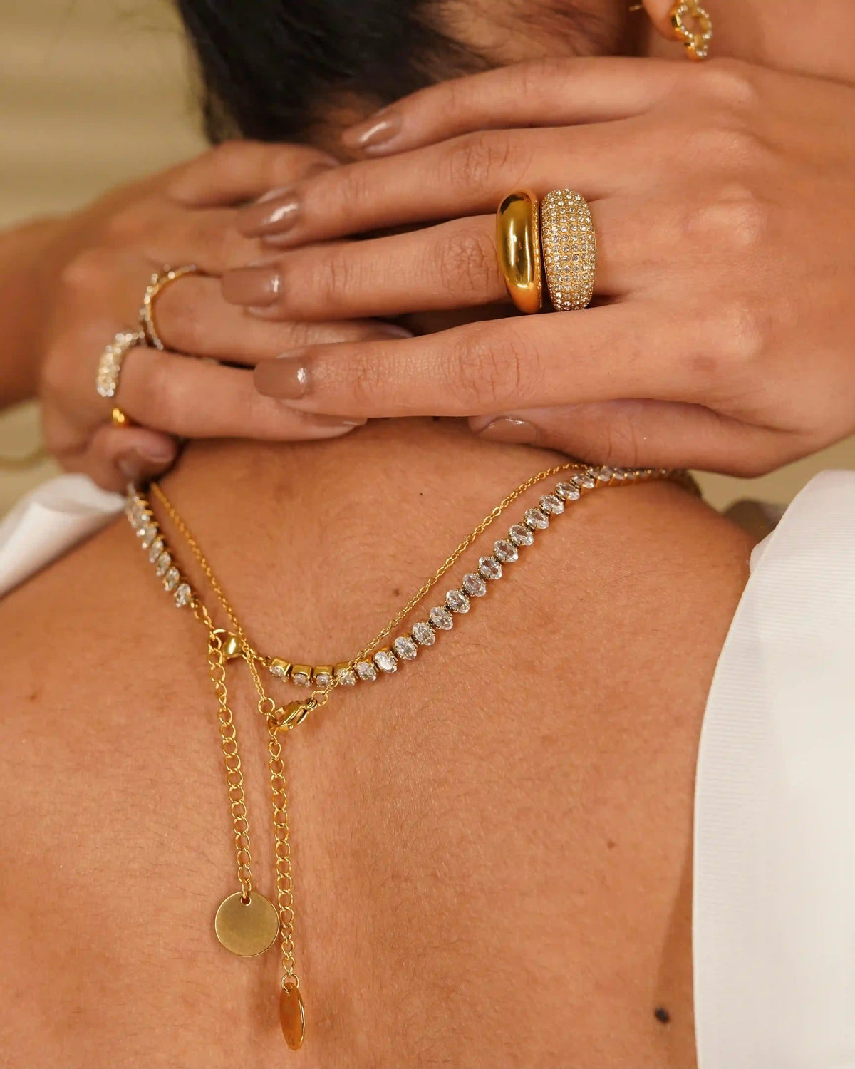 Back view of girl wearing linea tennis necklace with gold plated neckchain both hands above neck wearing 18k gold plated rings studded with zircons.