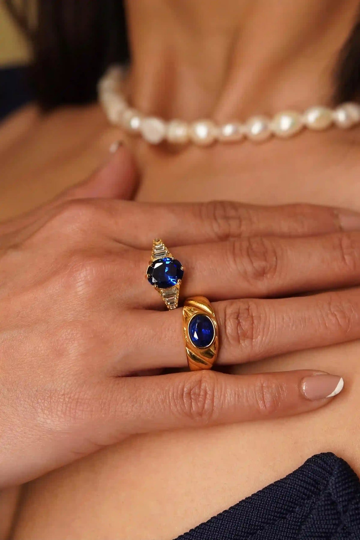 Close-up of a woman's hand wearing zepher sapphire ring and heirloom croissant sapphire ring having royal blue gemstones, with a pearl necklace in the background.