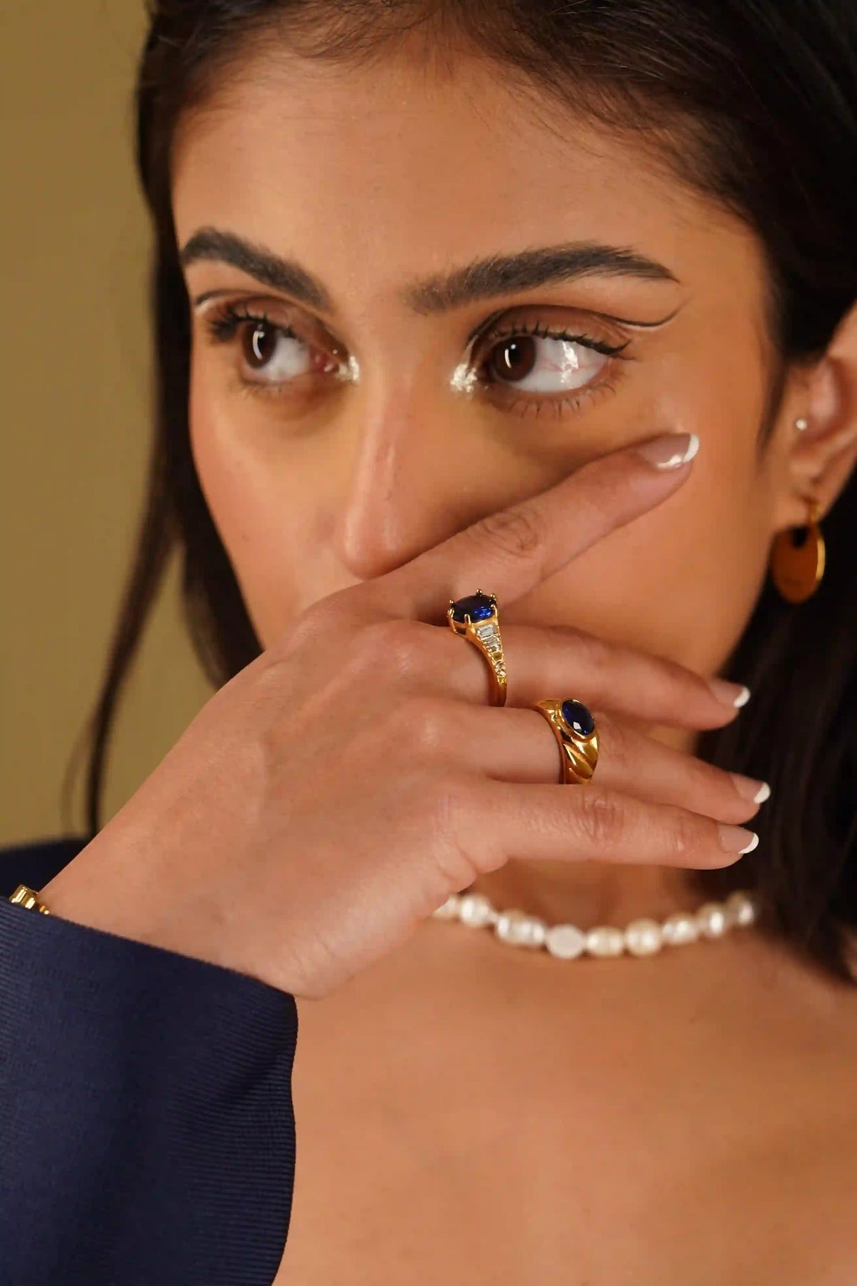 Close-up of a woman's face covering with hand wearing zepher sapphire ring and heirloom croissant sapphire ring and a pearl necklace.