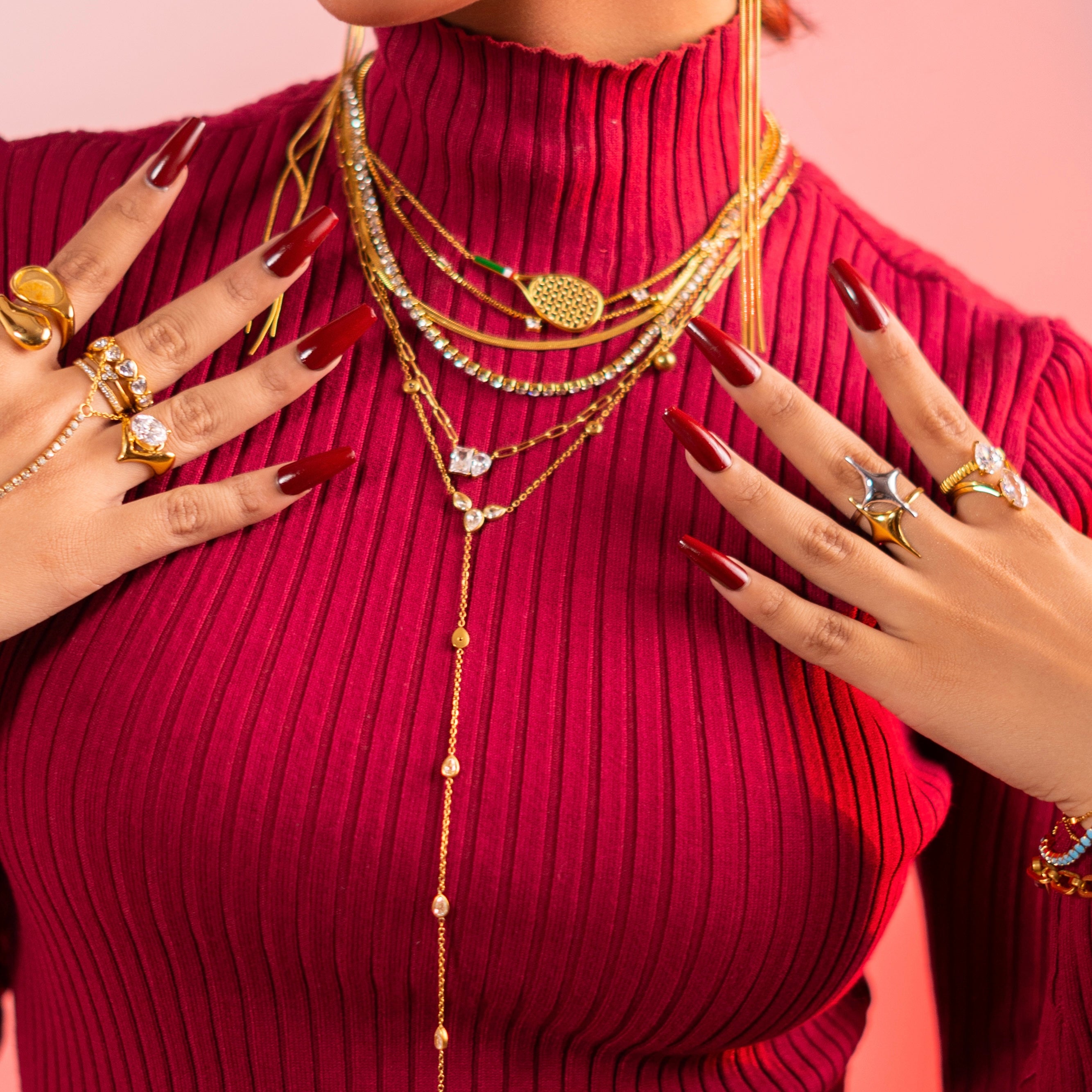 Girl wearing a red ribbed sweater with gold necklaces, earrings, and rings on a pink background