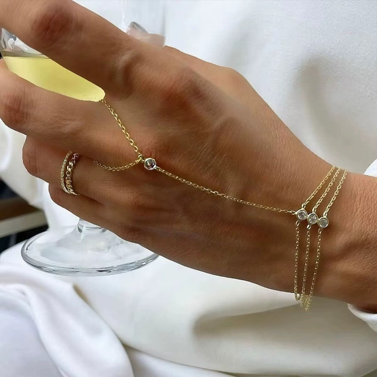 Hand wearing gold bracelets and rings with a glass of yellow liquid in the background