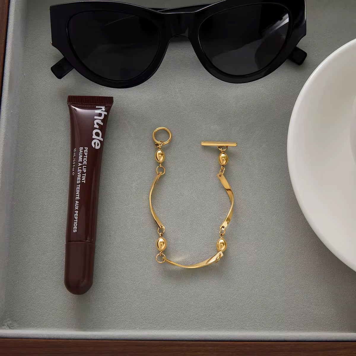 open bracelet and tube of makeup on a gray surface with sunglasses in the background