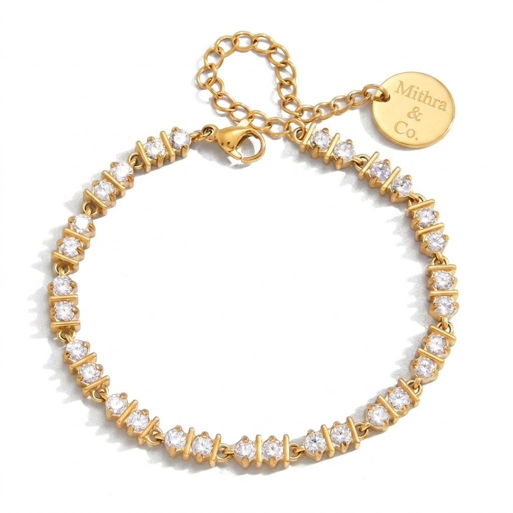 A gold tennis bracelet with clear round-cut stones in a spade setting, featuring a lobster clasp, extension chain, and a round gold tag engraved with 'Mithra & Co'.