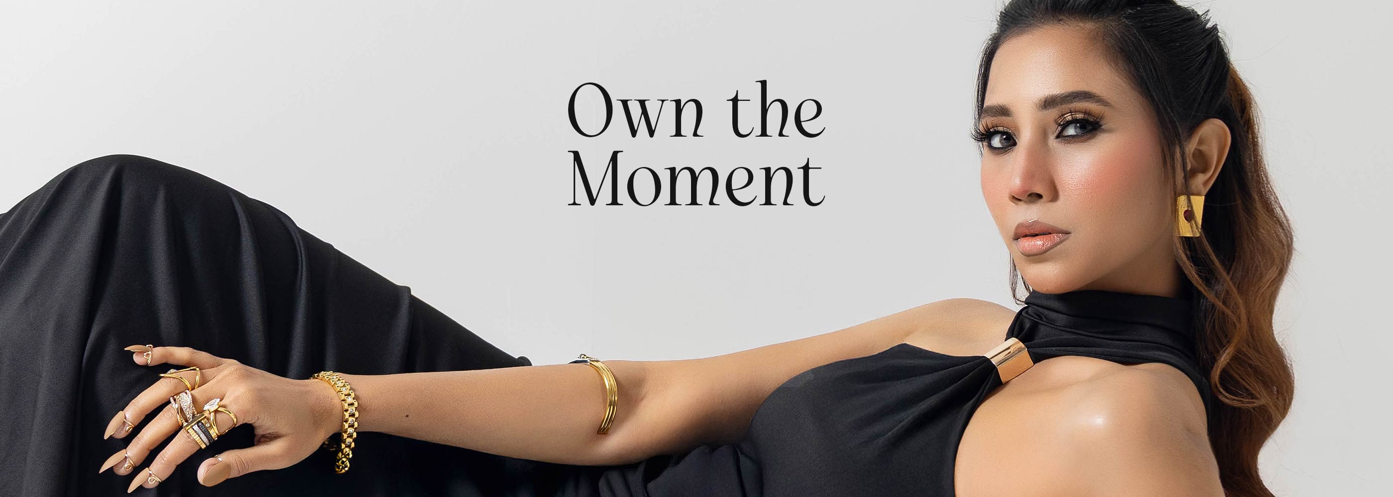 Woman in black outfit with gold plated jewelry, text 'Own the Moment' on a light gray background