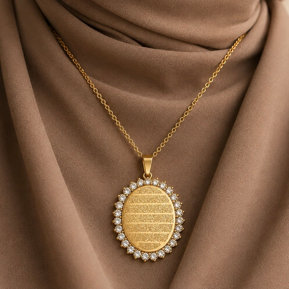 Ayatul Kursi Locket. A gold pendant necklace with a circular design, featuring a central gold oval with Arabic Calligraphy of Quranic Verse "Ayatul Kursi" and a border of small clear stones, displayed on draped brown fabric.