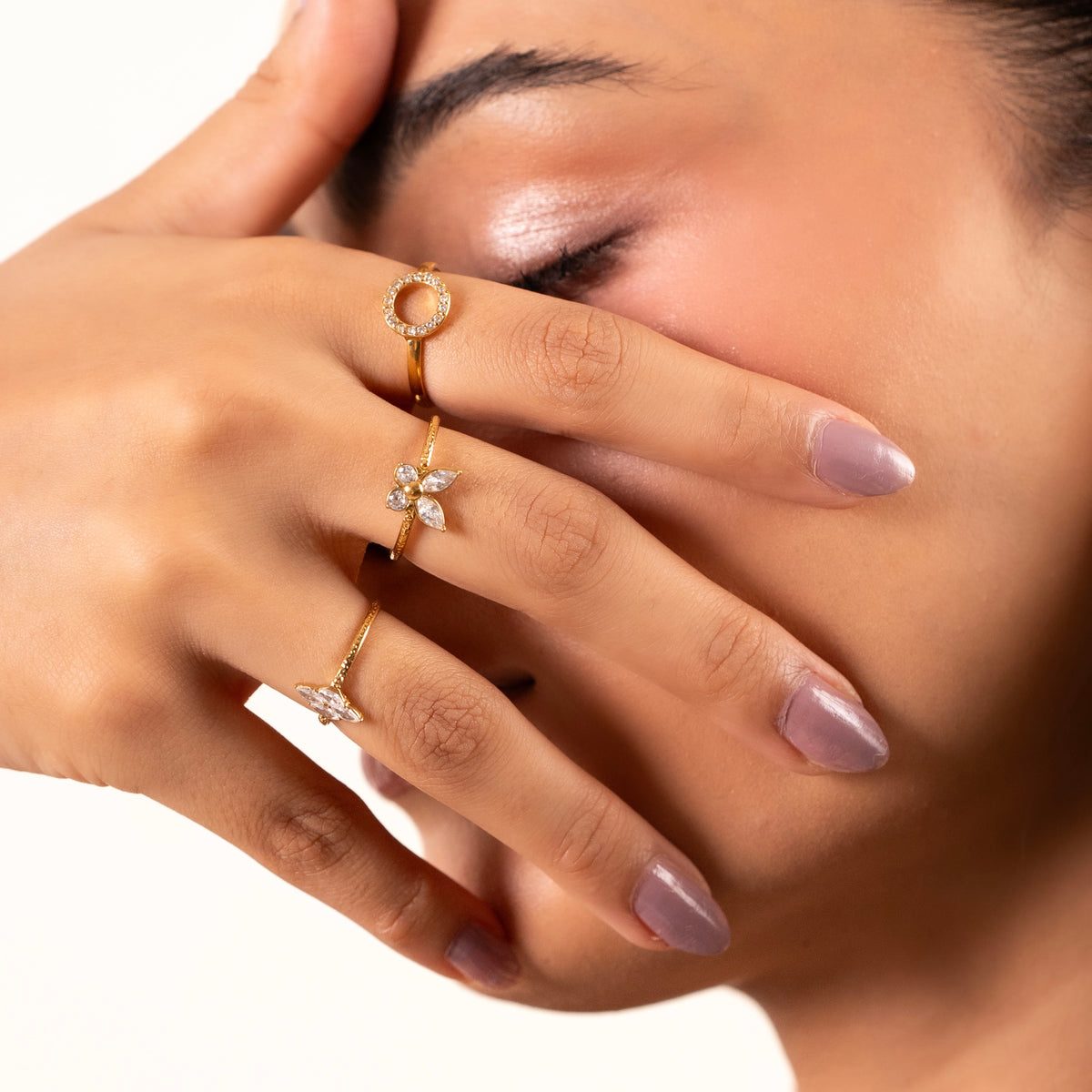 Bloom Stack Rings Set. A close-up lifestyle shot of a model's hand and face wearing three gold rings with clear gemstones, including a circular ring, a flower ring, and a small bird ring.