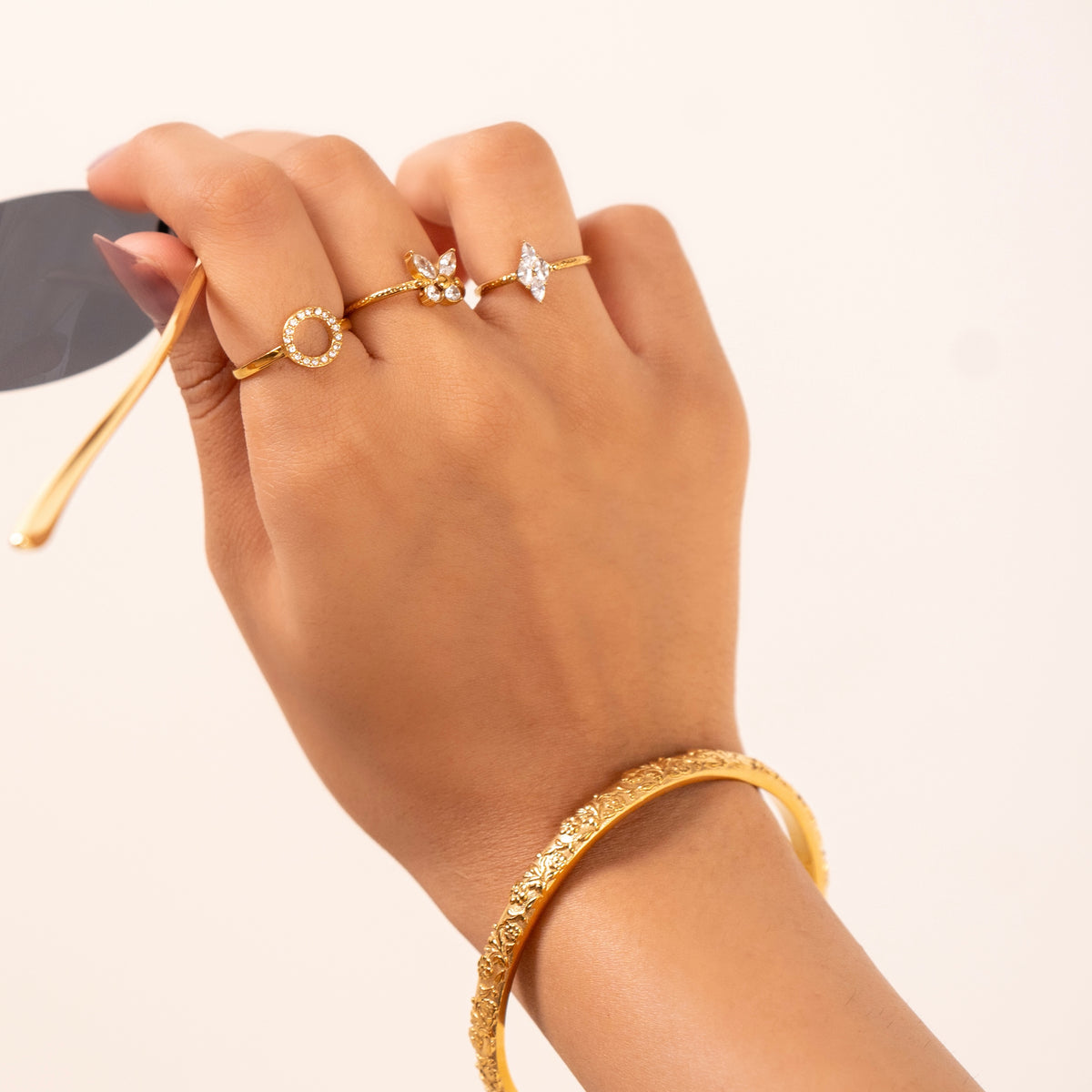 A close-up of a hand wearing multiple gold plated jewelry pieces, including a textured gold bangle, a bloom stack ring set including  ring with a circular pearl-like border, a ring with a small floral motif, and a ring with a star-shaped crystal. The hand is holding a spoon against a clean, off-white background.