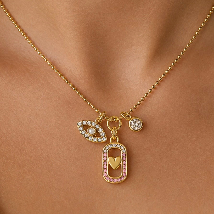 Gold charm necklace with an evil eye, a small round crystal bead, and a rectangular pendant with pink stones and a gold heart, shown on a model's neck.