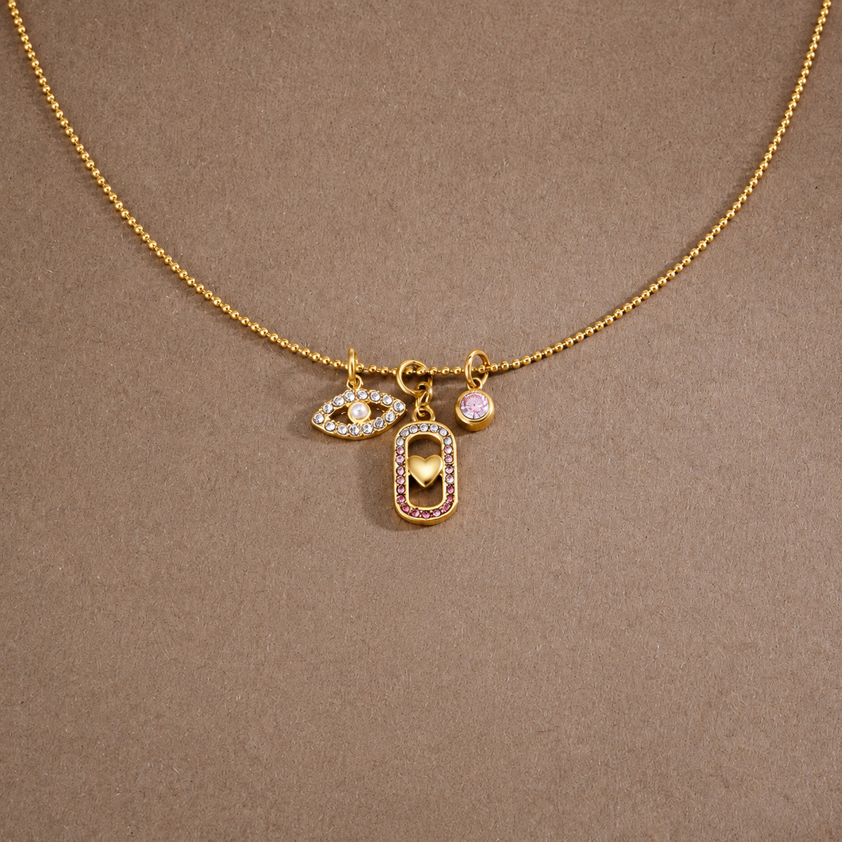 A gold chain necklace with three charms: an evil eye pendant, a rectangular pendant with a gold heart and pink crystal border, and a small round pink crystal charm, displayed on a textured brown background.