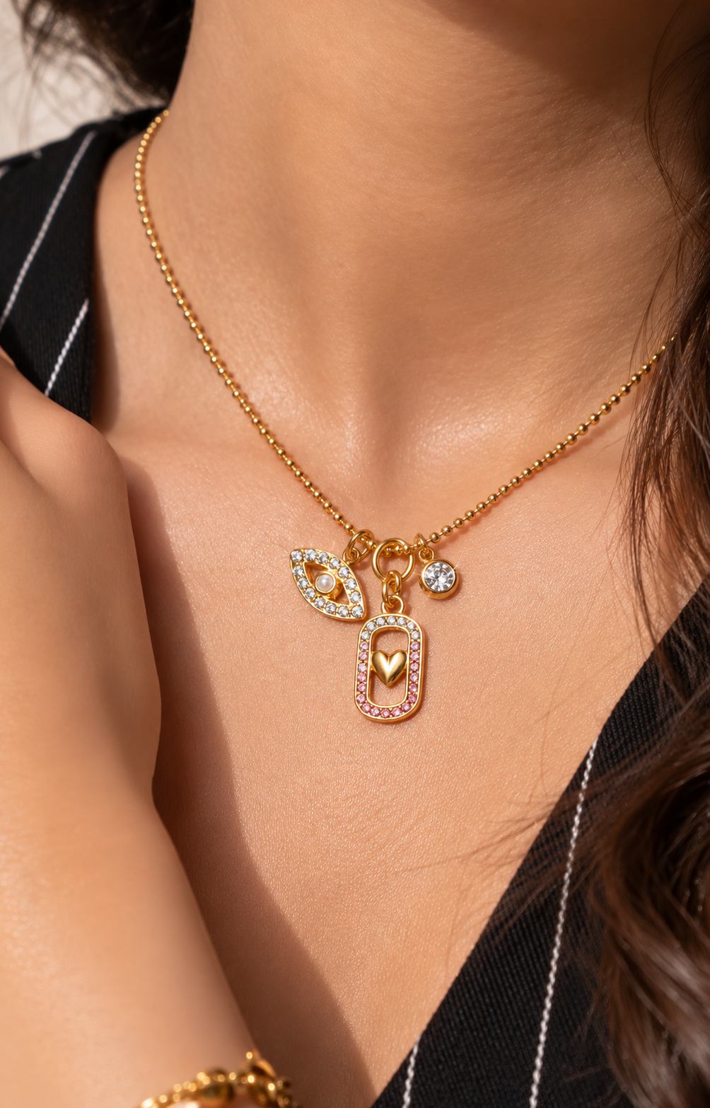 A close-up lifestyle shot of a model wearing a delicate gold chain necklace with a multi-charm pendant. The pendant features a small gold heart, a leaf-shaped charm set with clear crystals, and a small round clear stone, all resting on the model's chest.