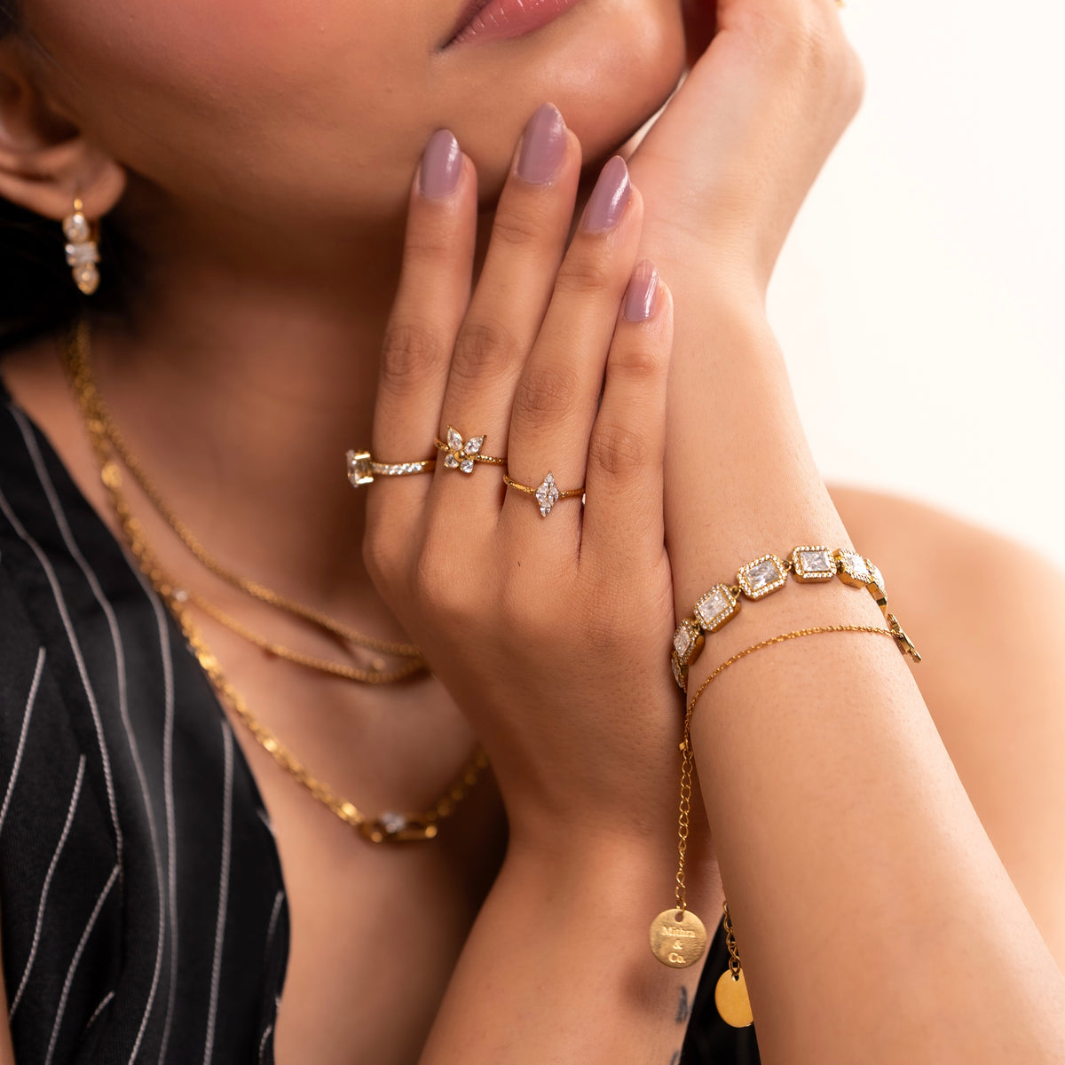 A close-up lifestyle shot of a model wearing a coordinated collection of gold plated jewelry, including a floral ring, a diamond-patterned ring, layered necklaces, emerald cut tennis bracelet, and a delicate chain bracelet with a round charm.