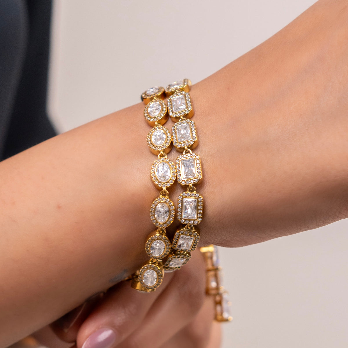 A close-up of a gold plated emerald cut tennis bracelet with multiple rows of round and rectangular clear gemstones worn on a person's wrist.