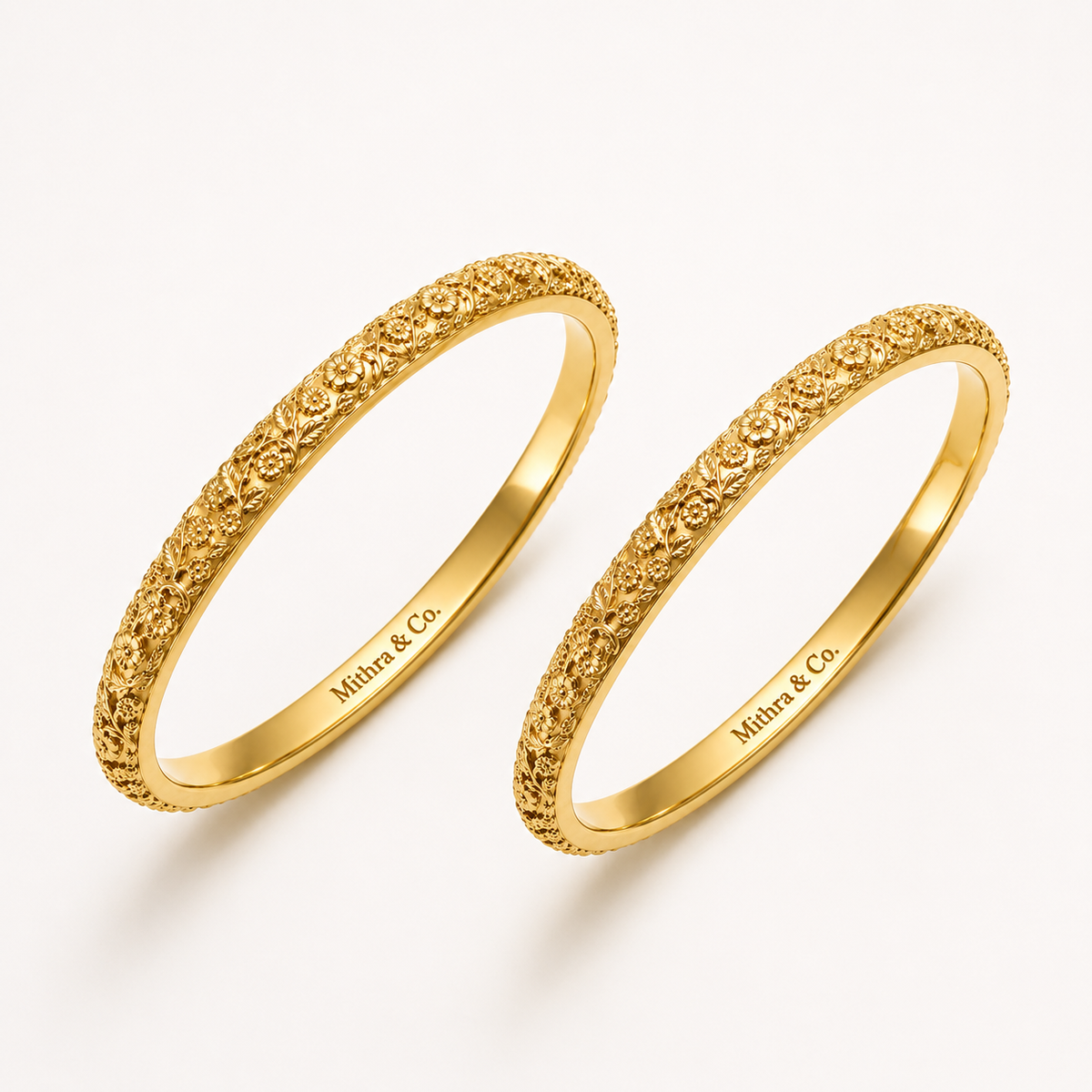 Two gold plated heritage bangle bracelets with textured floral detailing on the outer bands, displayed on a clean off-white background. The inner band is engraved with the brand name 'Mithra & Co.
