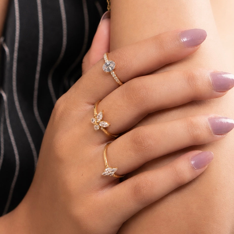 Three gold rings with clear gemstones in different floral and leaf designs, shown on a person's fingers. The person is wearing a black and white pinstripe top.