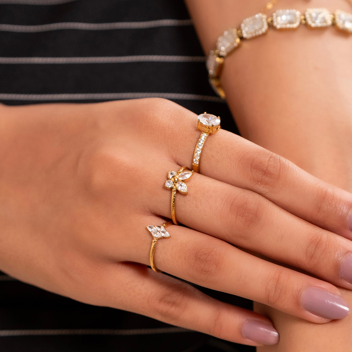 A close-up of a hand wearing three gold rings with clear gemstones, including butterfly motifs and a oval pavé band, alongside a matching gold bracelet.