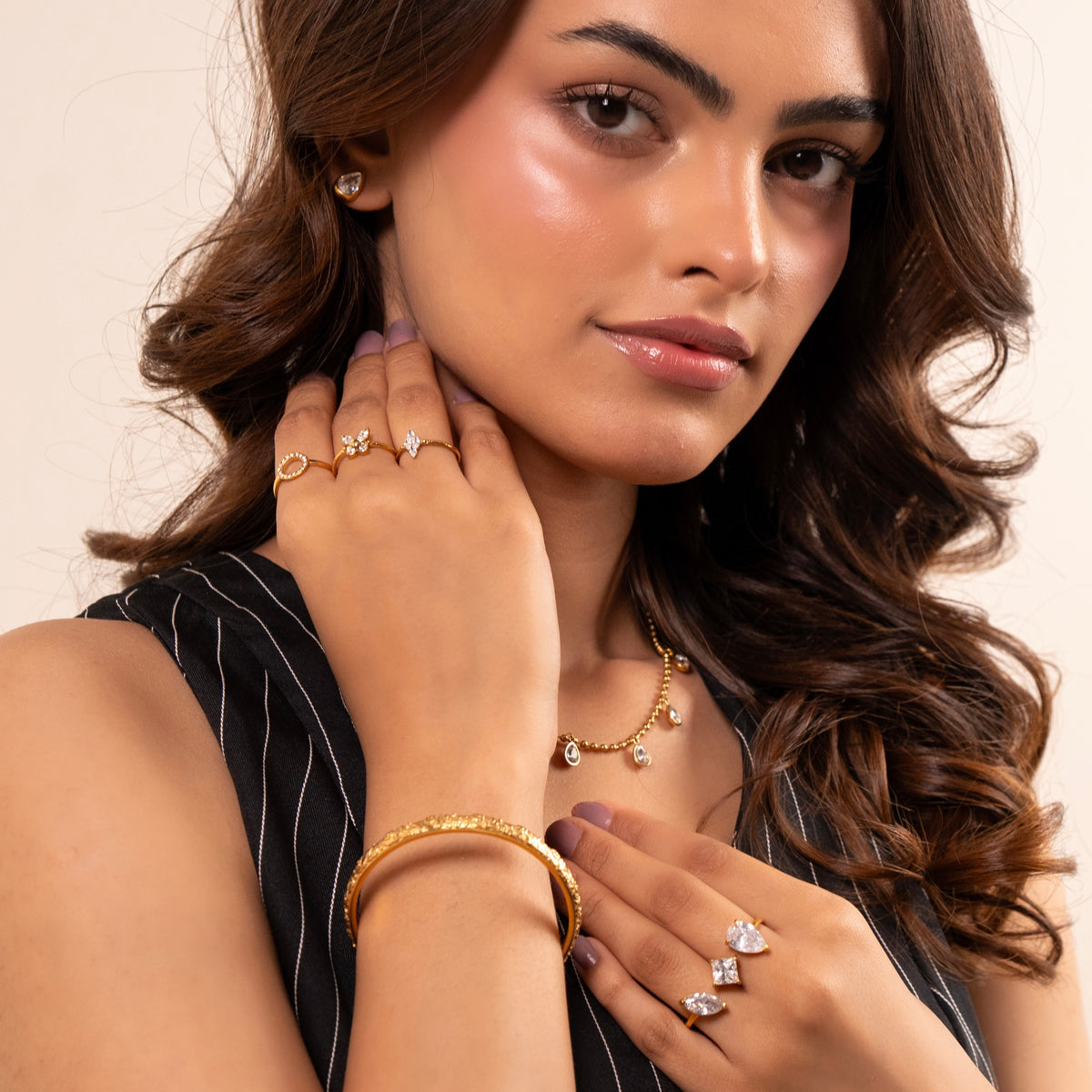 A close-up lifestyle shot of a woman wearing a gold teardrop jewelry set, including a textured gold bangle, delicate gold necklace with clear stones, and multiple gold rings, against a warm beige background.