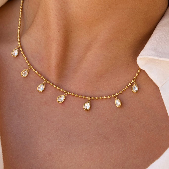 A gold chain necklace with seven small teardrop-shaped clear stones, worn on a model's neck.
