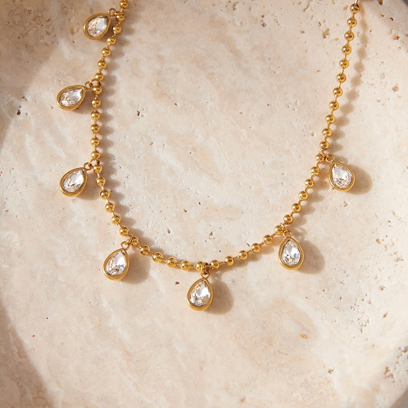 A gold beaded necklace with seven teardrop-shaped clear gemstones, displayed on a textured beige stone surface.