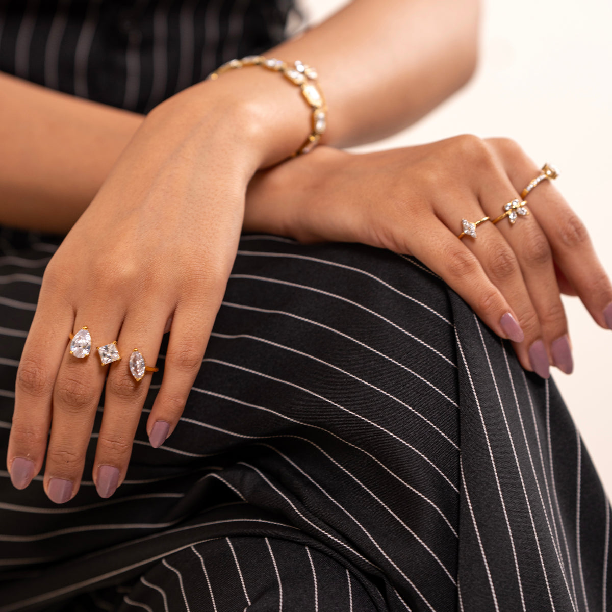 A close-up lifestyle shot of a person's hands and lap wearing multiple gold rings with clear gemstones, a gold bracelet, and black pinstriped trousers.