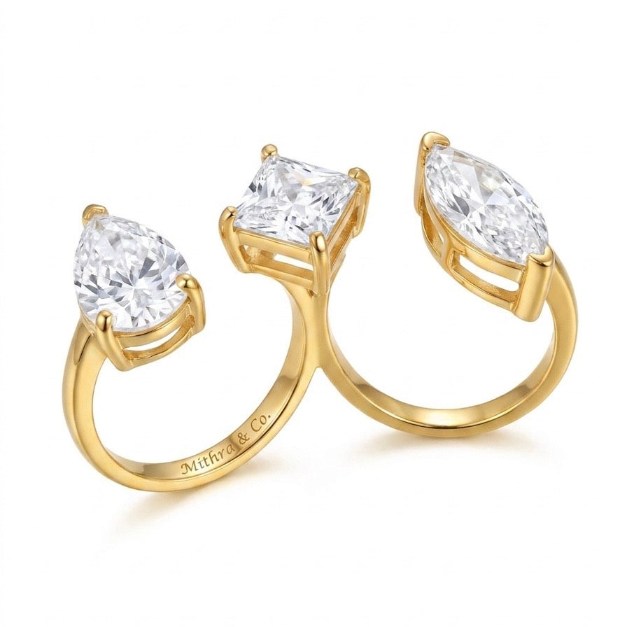 A gold dual finger ring with three gemstones in a connected cluster design: a pear-cut diamond on the left, a square princess-cut diamond in the center, and a marquise-cut diamond on the right. The inner band is engraved with 'Mithra & Co.