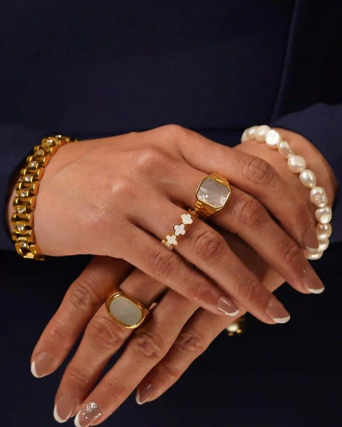 Closeup of a womans both hands wearing clover white band ring with glossy enamel design stacked with other gemstone rings and bracelets in a navy blue apparel bg.