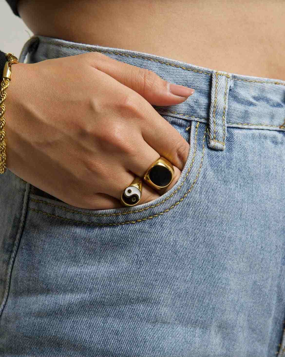a Hand Wearing gold-plated ramsey signet ring and yin and yan ring and twist chain bracelet casually placed in the front pocket of blue denim jeans.