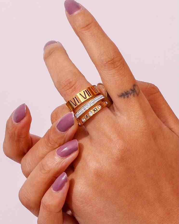Hand with triple layer 3 trilogy stacked ring with Roman Numerals and purple nail polish on a light pink background