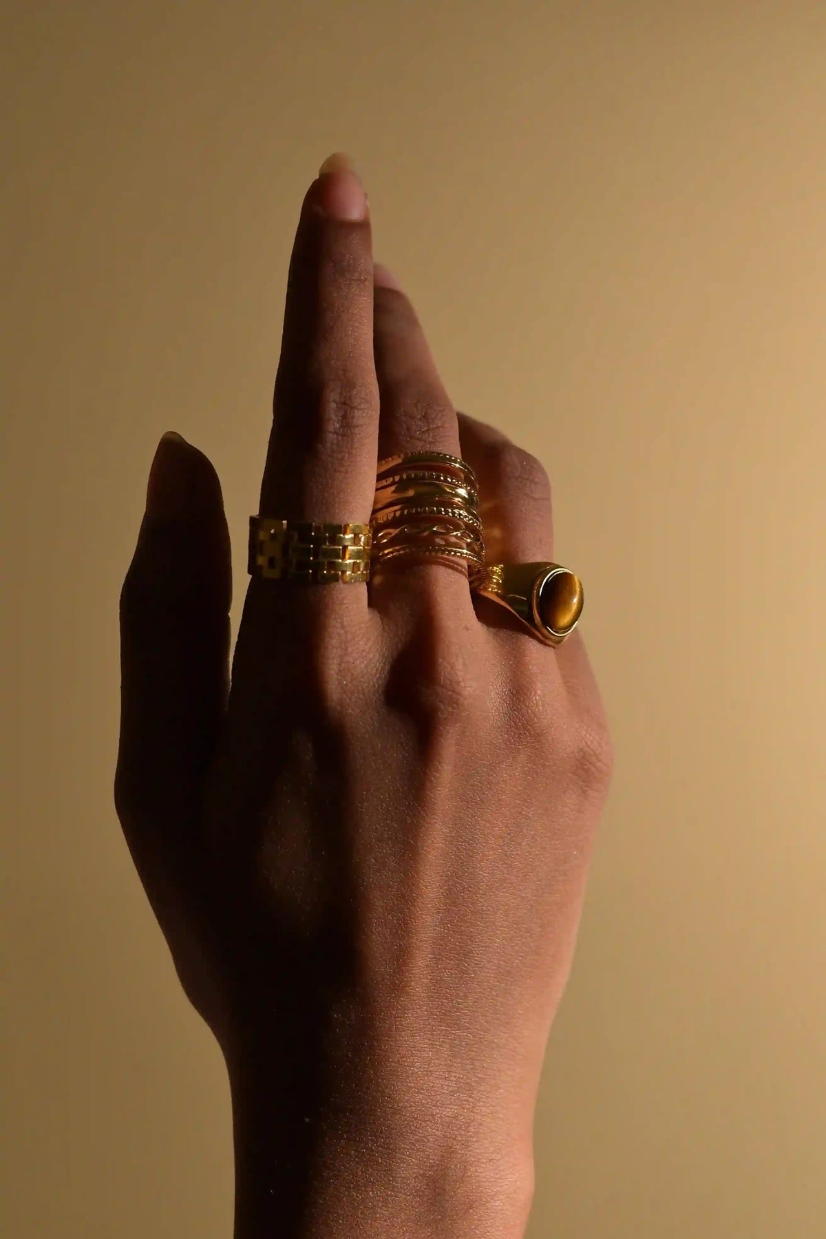 Closeup dark view of woman wearing 3 18k Gold Plated rings including mesh ring in index finger salento ring in middle finger and tiger's eye stone ring in ring finger on a pale bg.