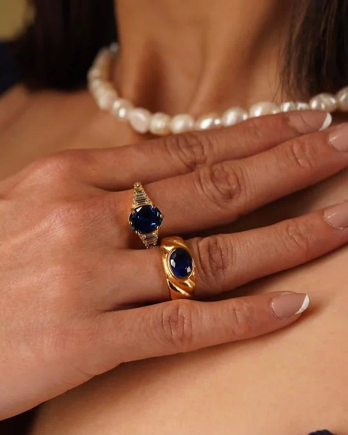 Close-up of a girl's hand wearing a zepher sapphire ring with Heirloom Croissant Sapphire Ring Both having royal blue gemstones, and a pearl necklace in the background.